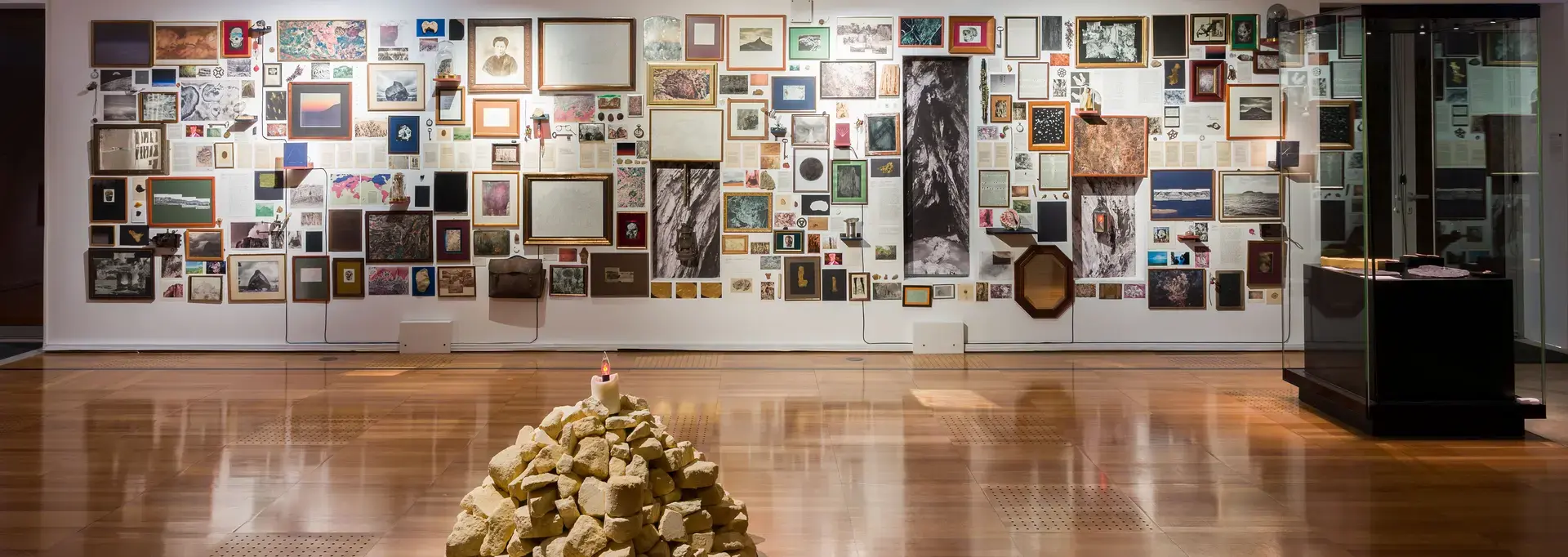 Vue d'ensemble d'une exposition d'Aurélien Mauplot présentée en 2025. Au premier plan, figure un amas de cailloux beiges surmonté d'une bougie allumée. À l'arrière-plan, un mur est tapissé de cadres et photos divers. Cet agencement retrace un cheminement des images à travers le temps, de la Préhistoire à nos jours.