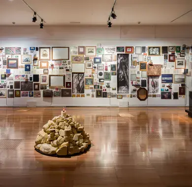 Vue d'ensemble d'une exposition d'Aurélien Mauplot présentée en 2025. Au premier plan, figure un amas de cailloux beiges surmonté d'une bougie allumée. À l'arrière-plan, un mur est tapissé de cadres et photos divers. Cet agencement retrace un cheminement des images à travers le temps, de la Préhistoire à nos jours.