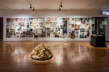 Vue d'ensemble d'une exposition d'Aurélien Mauplot présentée en 2025. Au premier plan, figure un amas de cailloux beiges surmonté d'une bougie allumée. À l'arrière-plan, un mur est tapissé de cadres et photos divers. Cet agencement retrace un cheminement des images à travers le temps, de la Préhistoire à nos jours.